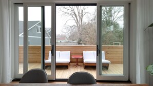 A room with a sliding glass door leading to an outdoor deck with white cushioned seating and a small round table. - Modernist Masterpiece: 'Simply the Best' (Durham)
