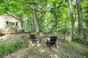 Une terrasse en pierre avec des fauteuils Adirondack, entourée d’une zone boisée où s’élèvent de grands arbres.