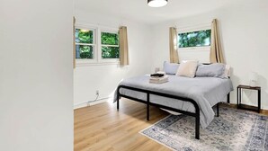 A bedroom with a bed, two windows with curtains, a bedside table, and a rug. - Boiler Bungalow - entire 3(King)Bed home! (West Lafayette)