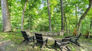 A forested area with a stone wall, a fire pit, and four wooden chairs. - Boiler Bungalow - entire 3(King)Bed home! (West Lafayette)
