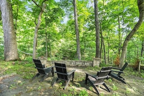 A forested area with a stone wall, a fire pit, and four Adirondack chairs.