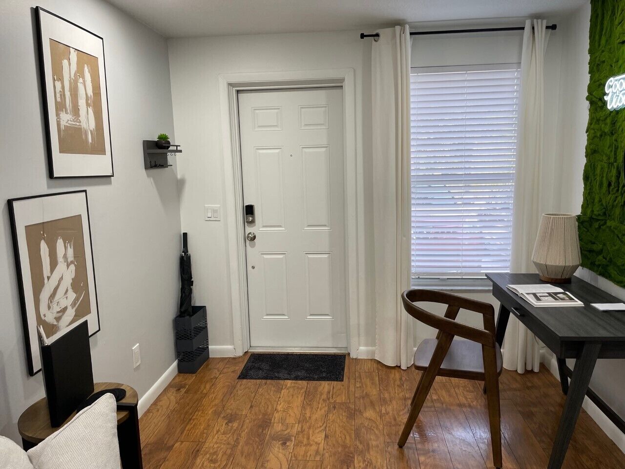A modern living room with a wooden floor, a white door, a black chair, a small table with a lamp, and framed artwork on the wall.