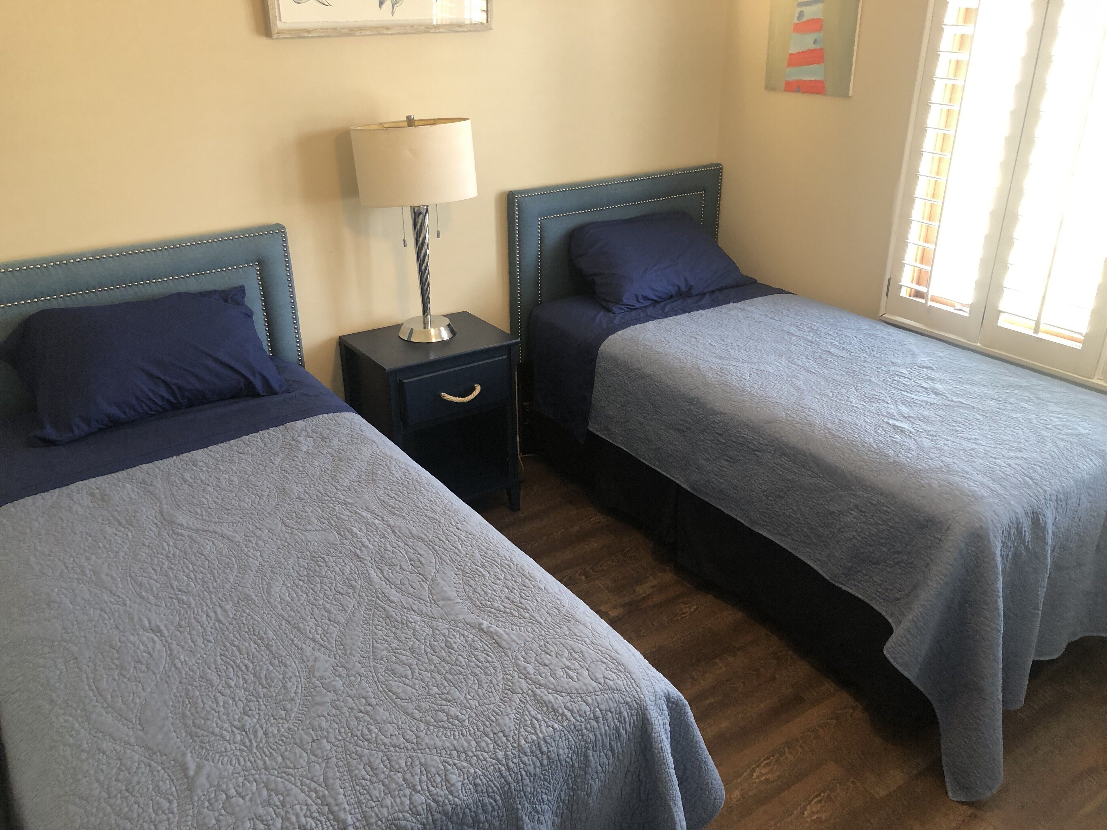 A room with two beds, each with a headboard and a bedside table with a lamp.