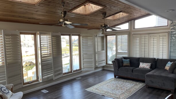 A spacious living room with a dark wood floor, a gray sofa, and a large window with white shutters.