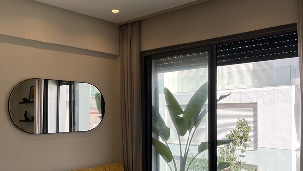Un salon moderne avec un canapé jaune, un miroir rond, une petite table et un balcon agrémenté d’une plante en pot.