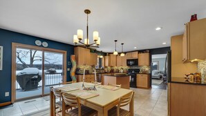 A modern kitchen with wooden cabinets, a central island, and a dining area with a table and chairs. - Chain of Lakes Waterfront Home (Central Lake)