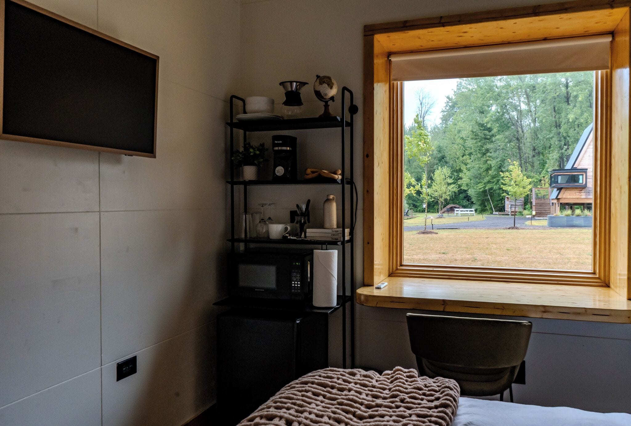 A bedroom with a bed, a wooden chair, a shelf with kitchen items, a window with a view of trees and a building, and a wall-mounted TV.