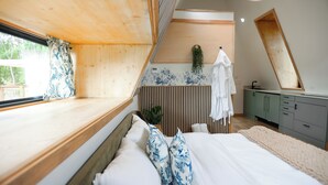 A bedroom with a sloped ceiling, a large window, a bed with white bedding and blue pillows, a bedside table, a dresser, and a mirror. - Finger Lakes Treehouse & Spa (Sodus)