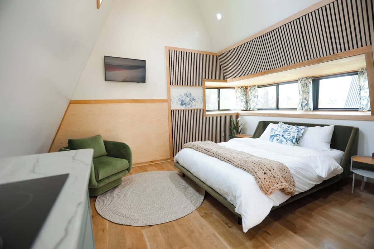 A modern bedroom with a bed, a green chair, a round rug, and a view of the outdoors through a window with curtains.