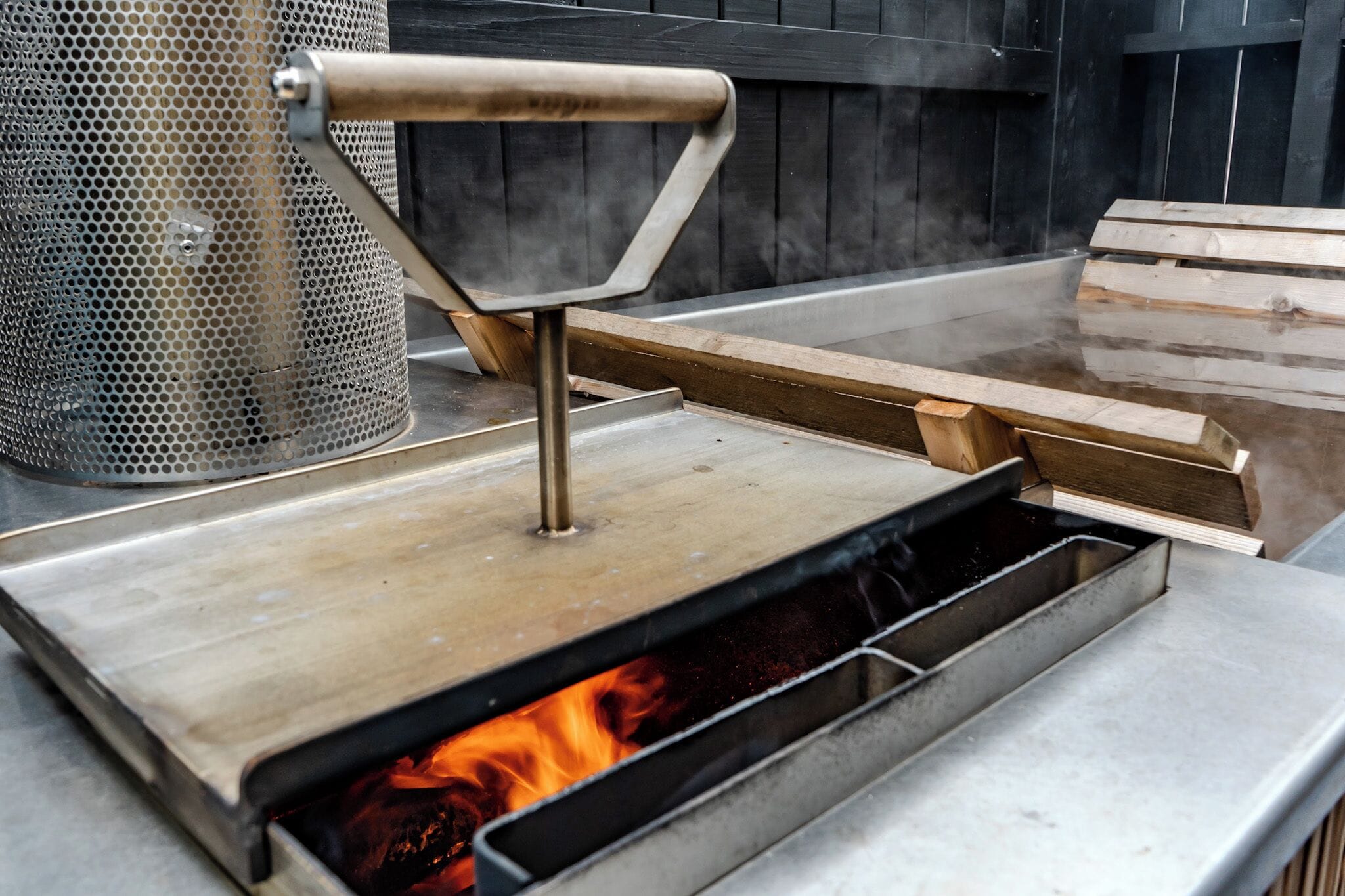A metal grill with a handle, a mesh container, and wooden planks on a flat surface.