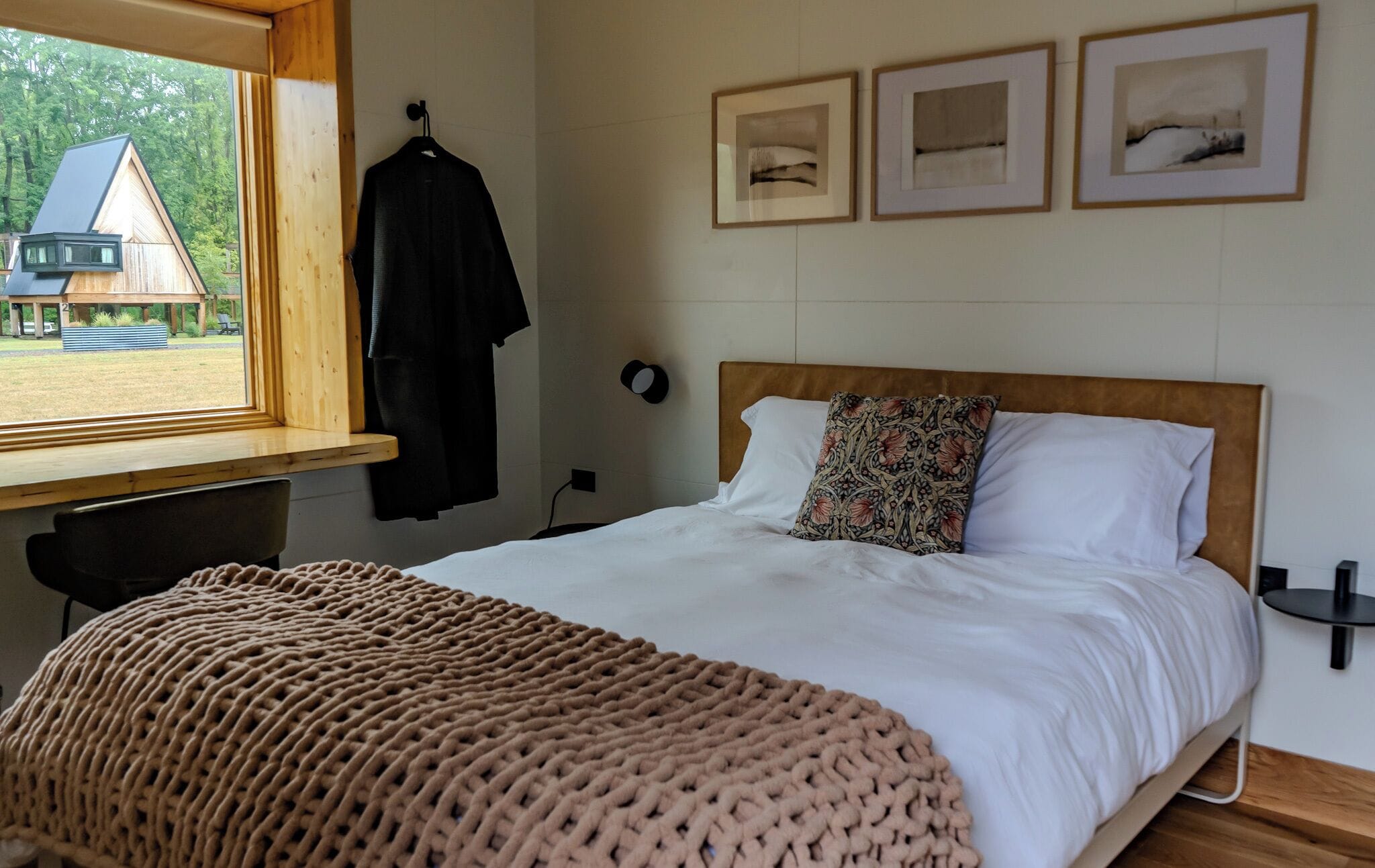 Un dormitorio con cama, ventana con vista a una casa, escritorio y silla.