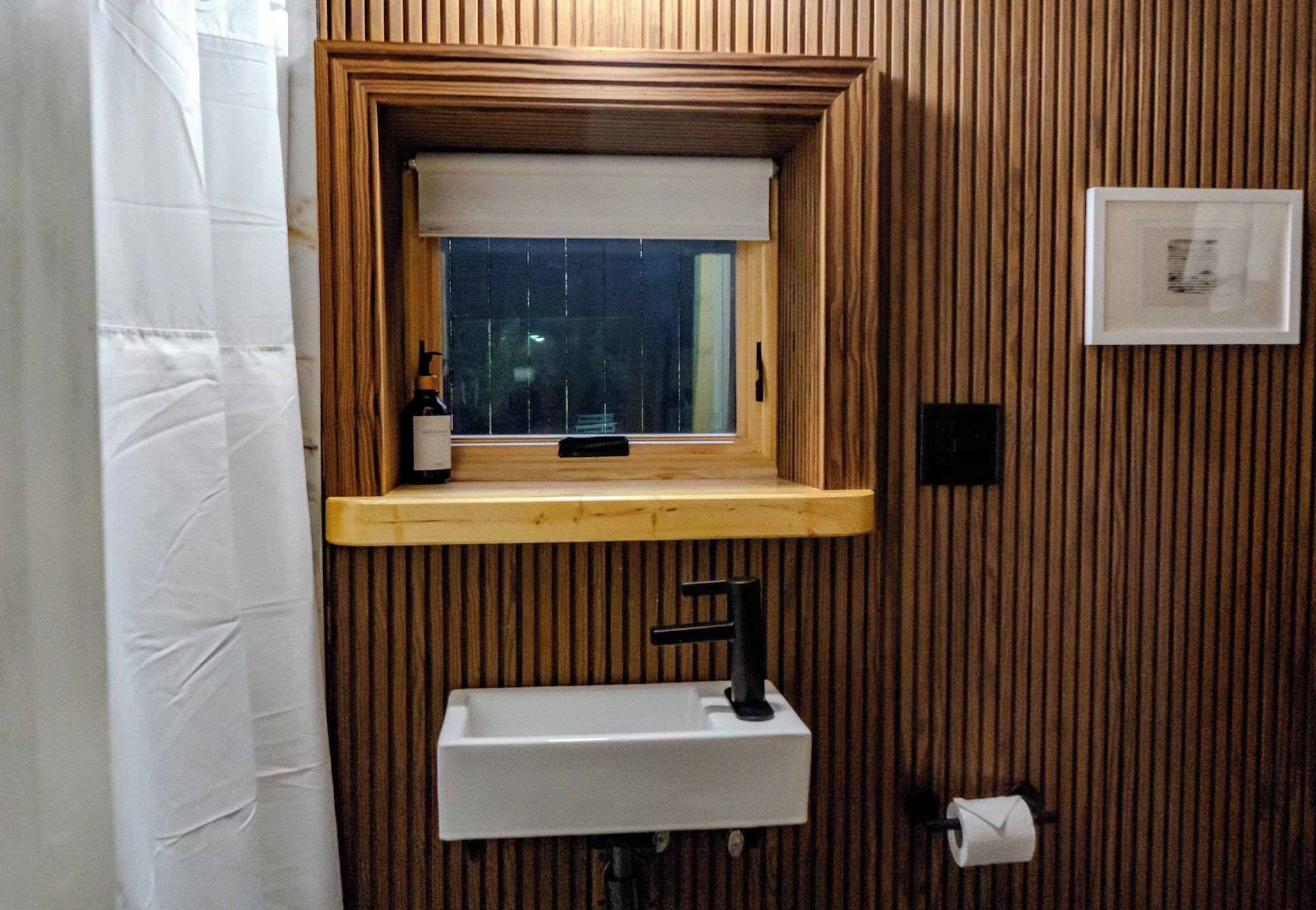 Um banheiro com parede de madeira, janela com persiana branca, pia com torneira preta e uma garrafa de shampoo no balcão.