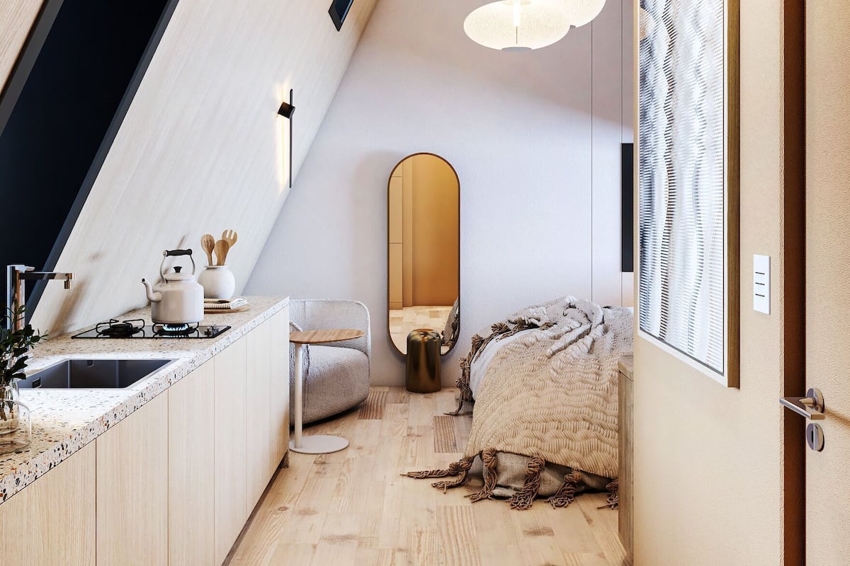 A modern loft bedroom with a sloped ceiling, a built-in kitchen with a sink and stove, a cozy seating area, and a large mirror.