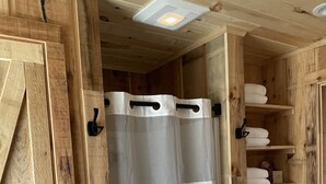 A wooden interior with a white shower curtain, wooden shelves with towels, and a metal basket. - Ridgeview Cottage (Lexington)
