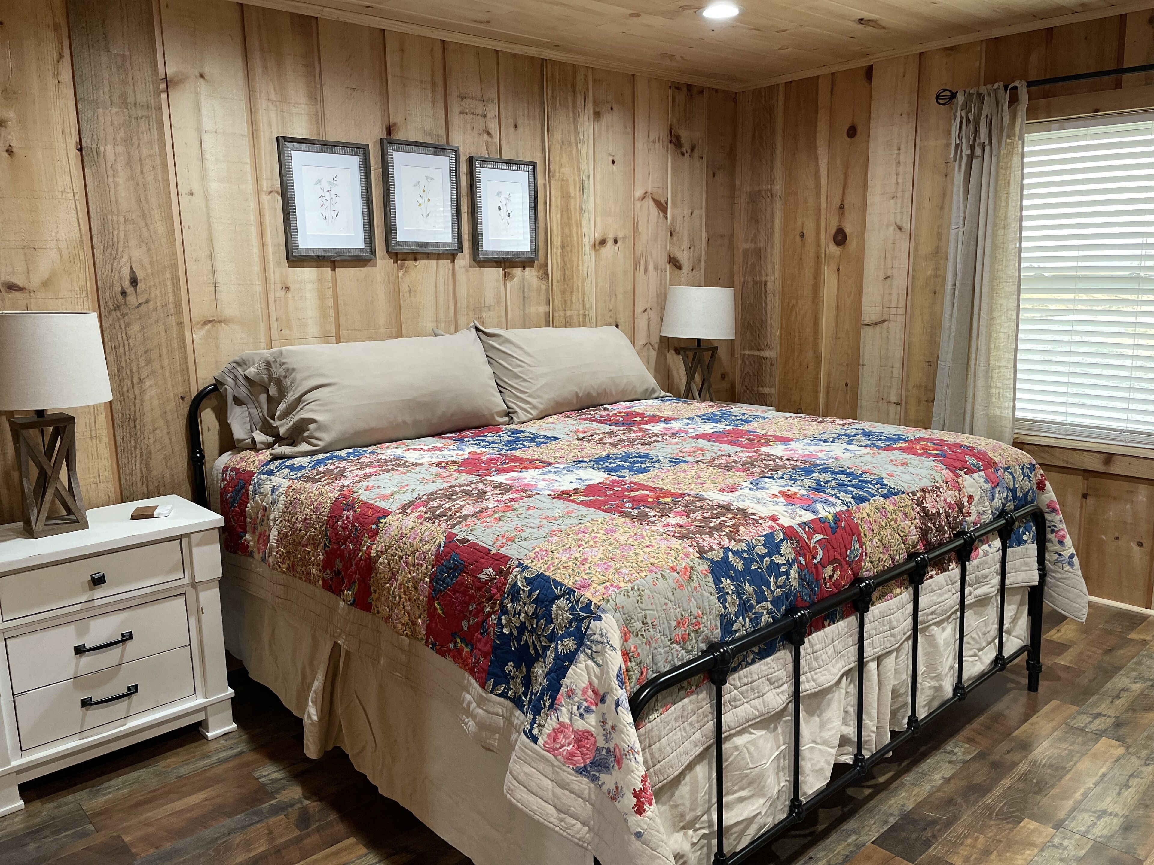 A bedroom with a wooden paneled wall, a bed with a floral quilt, two bedside lamps, a nightstand, and a window with blinds.