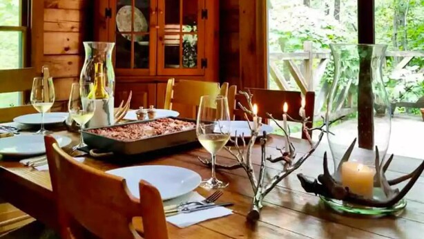A wooden cabin interior with a dining table set for two, a cabinet with glass doors, and a view of the forest through large windows.