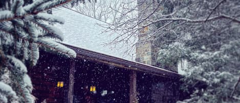 A wooden cabin with a snow-covered roof, surrounded by snow-laden trees and bushes.