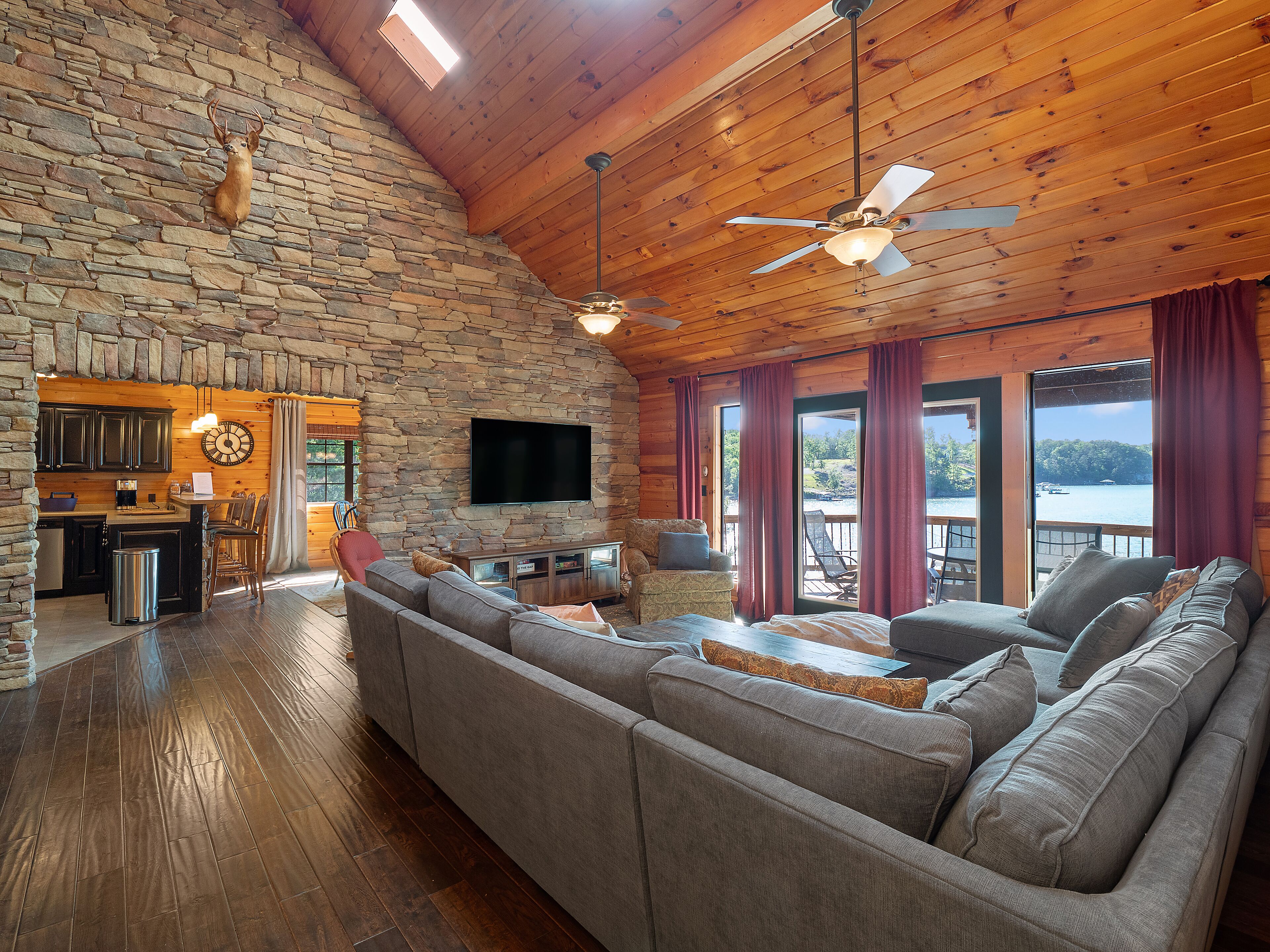 A cozy cabin interior with a stone wall, wooden ceiling, a large sofa, a flat-screen TV, and a view of a lake.