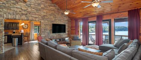 A cozy cabin interior with a stone wall, wooden ceiling, a large sofa, a flat-screen TV, and a view of a lake.