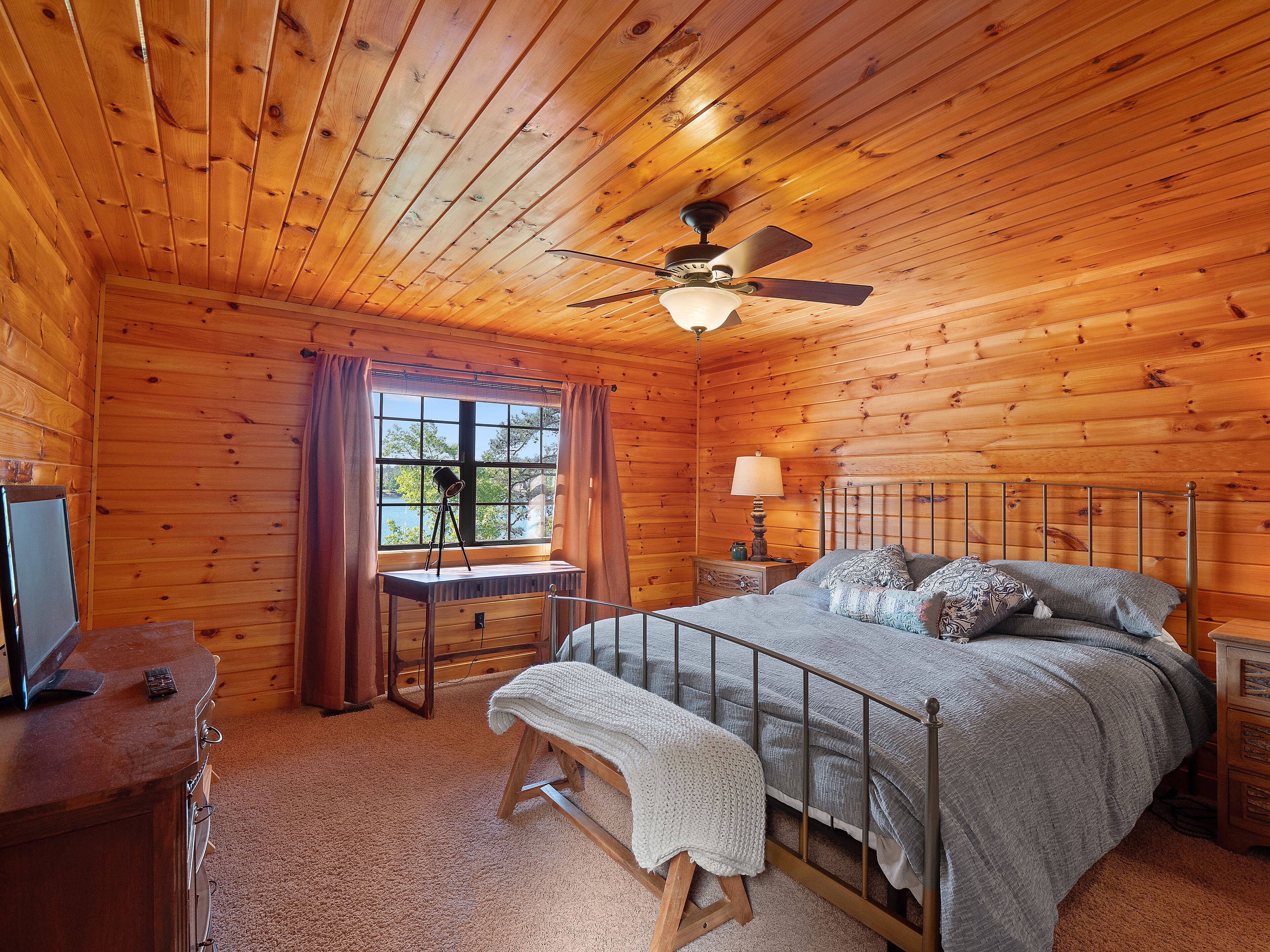Ein Schlafzimmer einer Holzhütte mit einem Bett, einem Schreibtisch, einem Stuhl, einer Lampe und einem Deckenventilator.