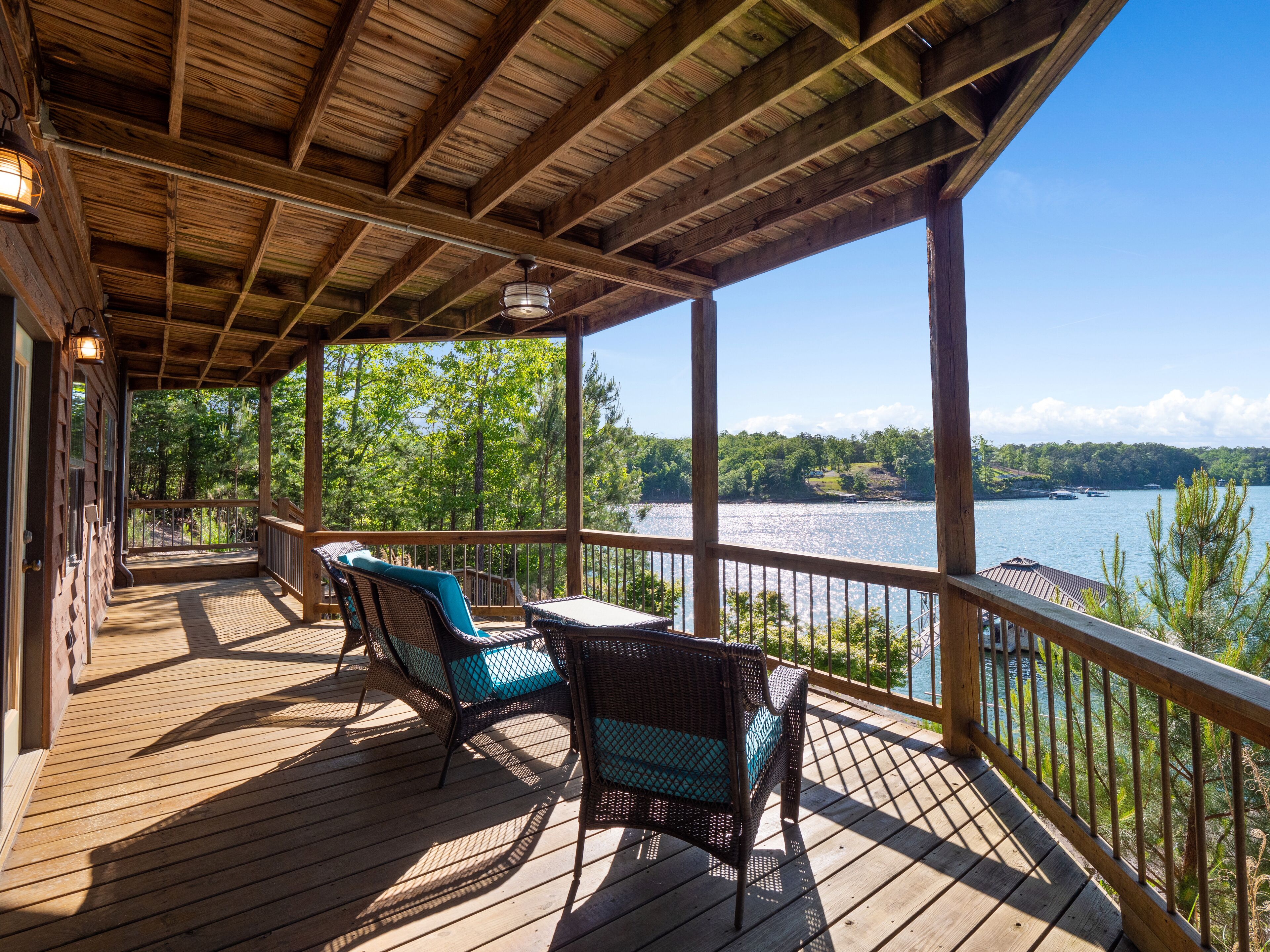 A wooden deck with wicker chairs and a table, overlooking a lake with boats and trees.
