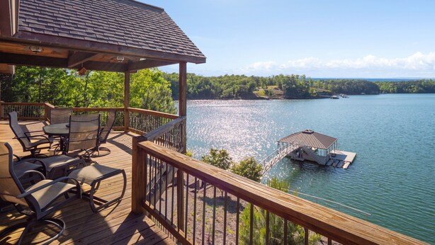 Una terraza de madera con mobiliario exterior que da a un lago, un muelle y un área boscosa.