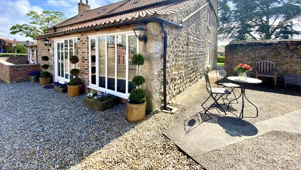 A stone cottage with a tiled roof, large windows, and a gravel patio with outdoor seating. - Grange Farm Lodge (Ripon)