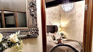 A bedroom with a large ornate mirror, a tufted headboard, and a bedside table with a vase of flowers. - Grange Farm Lodge (Ripon)