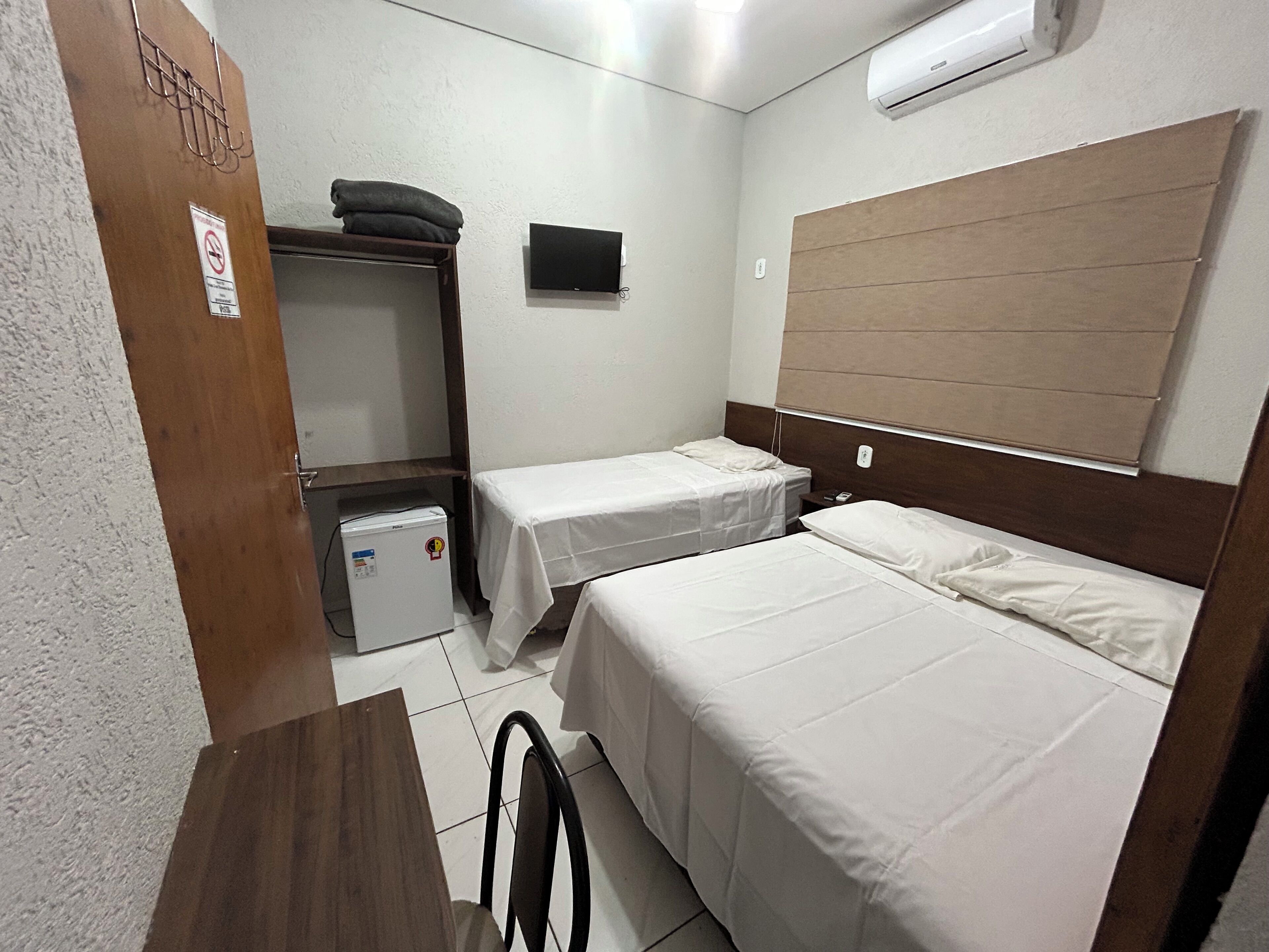 Quarto pequeno e limpo, com duas camas de solteiro, uma escrivaninha, um frigorífico e uma televisão na parede.