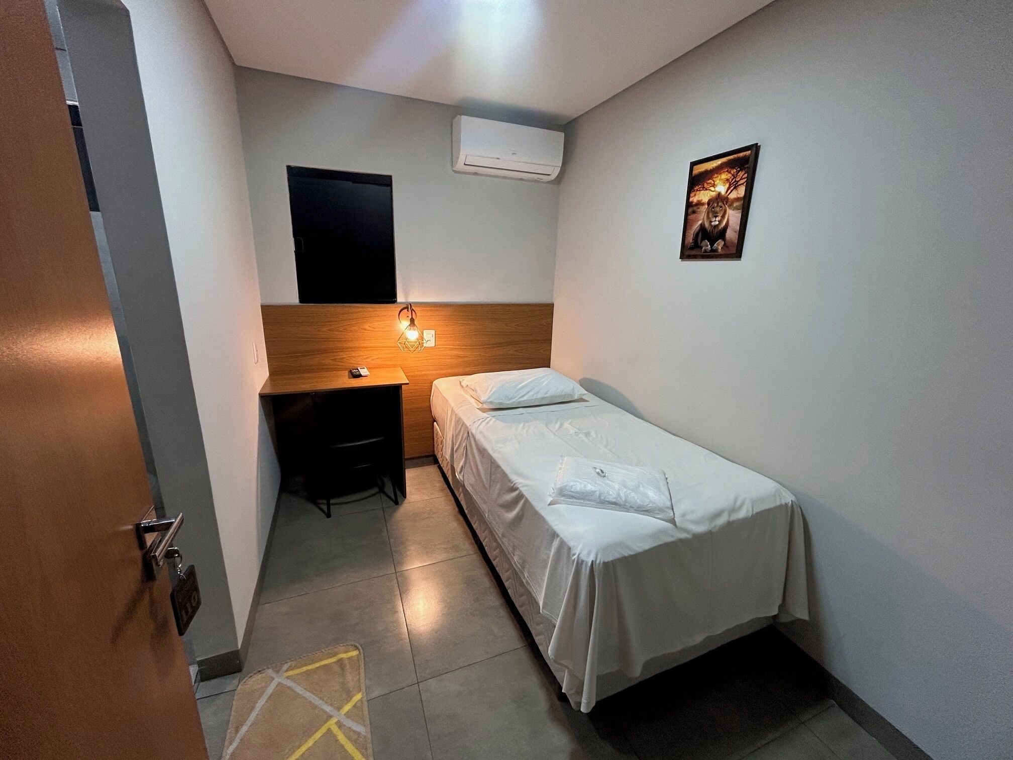Une petite pièce propre, avec un lit simple, un bureau et un climatiseur fixé au mur.