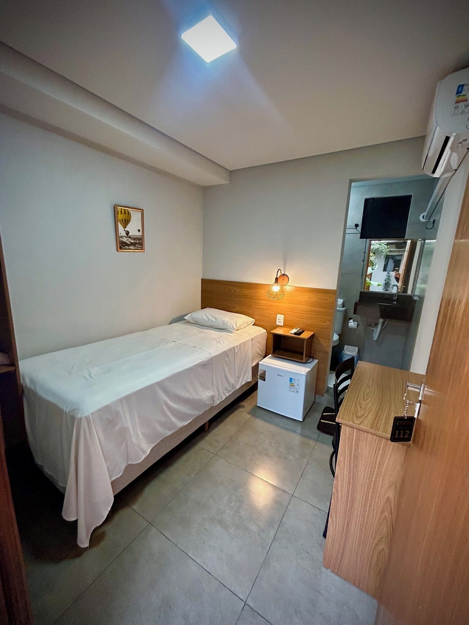 A small, neatly arranged room with a single bed, a bedside table, a small refrigerator, a wooden cabinet, and a wall-mounted TV.