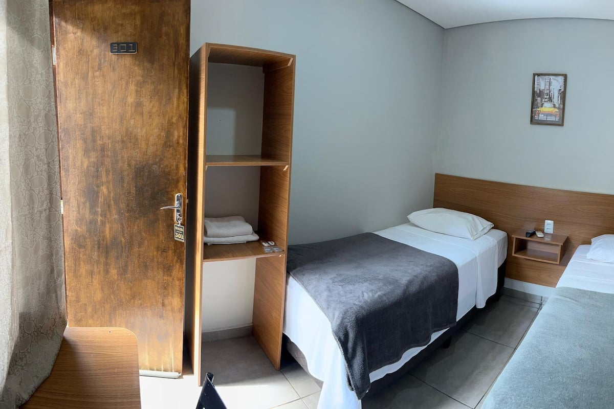 Een kleine hotelkamer met een houten garderobe, twee bedden, een nachtkastje en een aan de muur hangend schilderij.