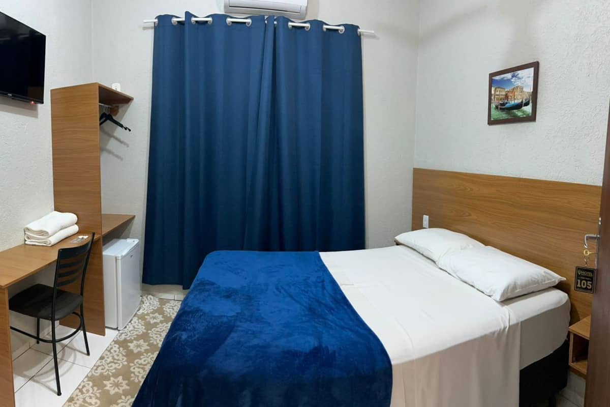Een kleine, opgeruimde kamer met een bed, een bureau, een stoel en een televisie aan de muur.