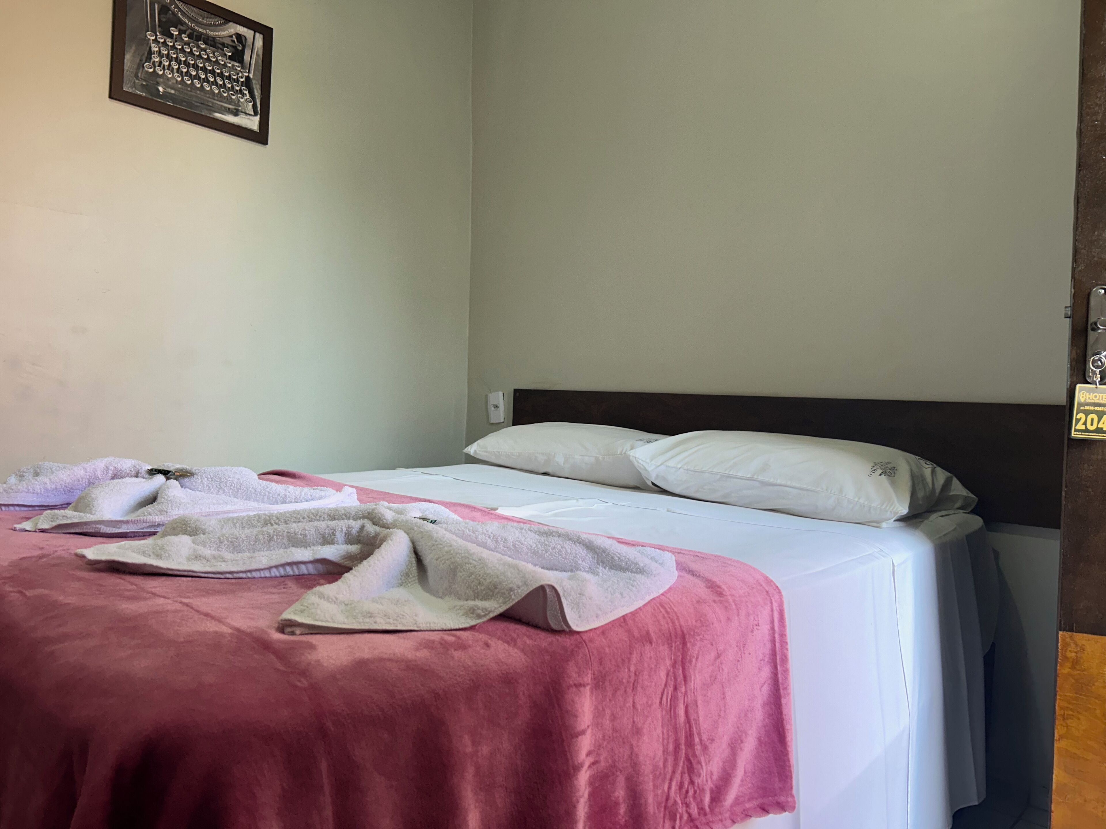 A single bed with a white mattress and two white pillows, a pink blanket, and a white towel.