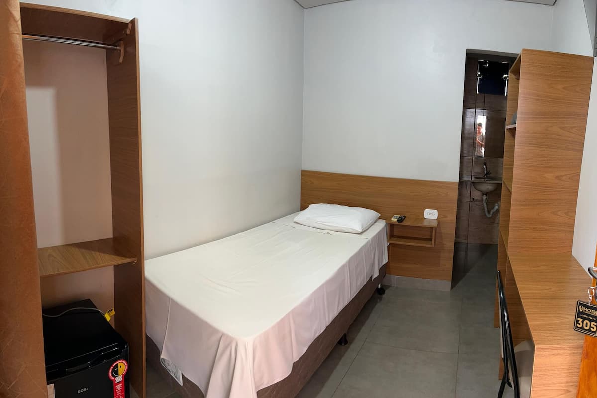 Een kleine, schone kamer met een eenpersoonsbed, een houten garderobe, een nachtkastje met telefoon en een badkamer met wastafel en spiegel.
