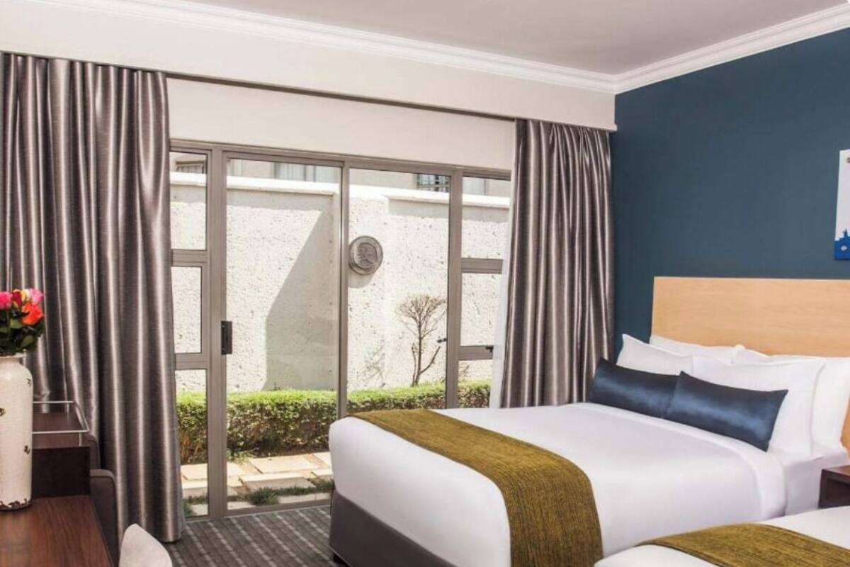 Quarto de hotel com duas camas, uma mesa de trabalho e vista para o exterior através de uma porta deslizante.