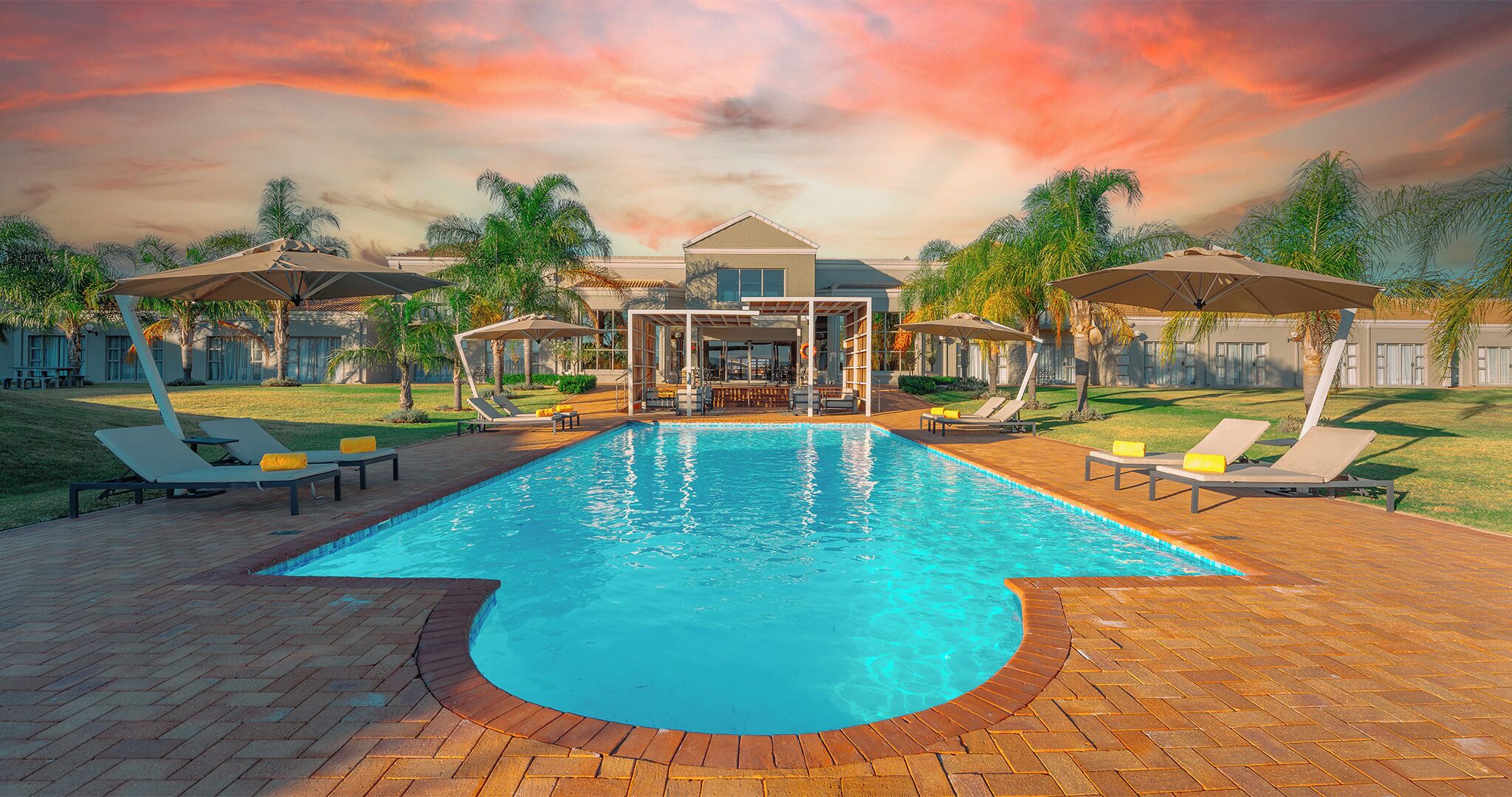 A rectangular swimming pool with a brick border, surrounded by lounge chairs and umbrellas, with a building and palm trees in the background.