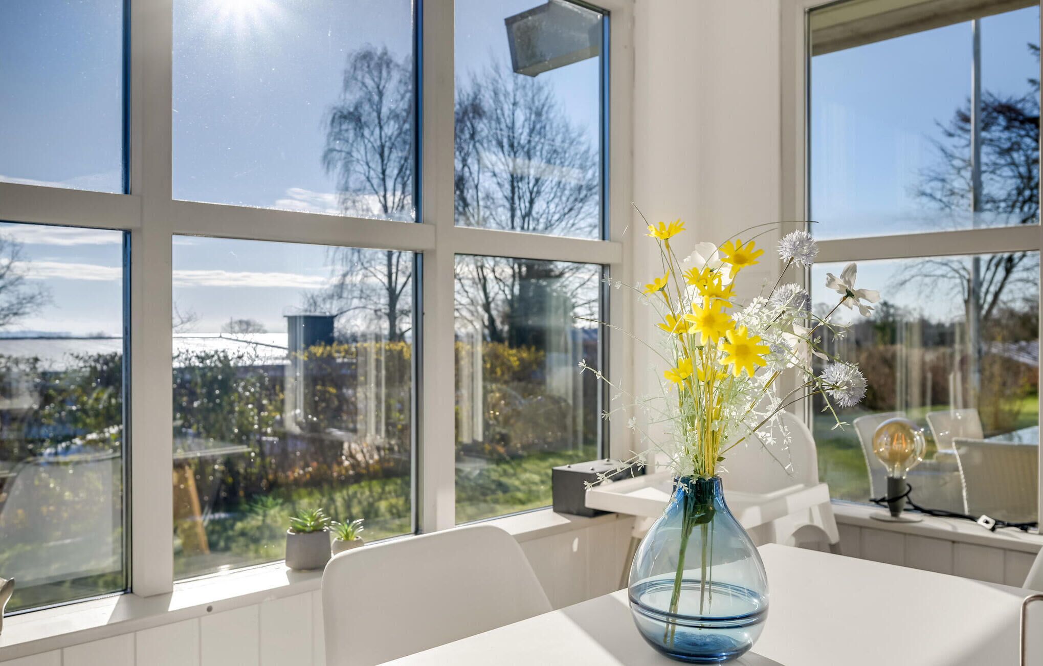 Una habitación con ventanas amplias, un florero con flores amarillas y vistas a un jardín y árboles.