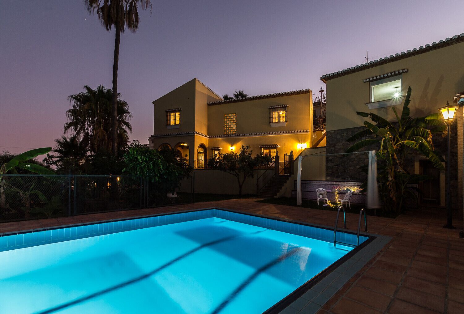 A swimming pool with a tiled deck, surrounded by a fenced area with palm trees and a building with multiple windows and a balcony.