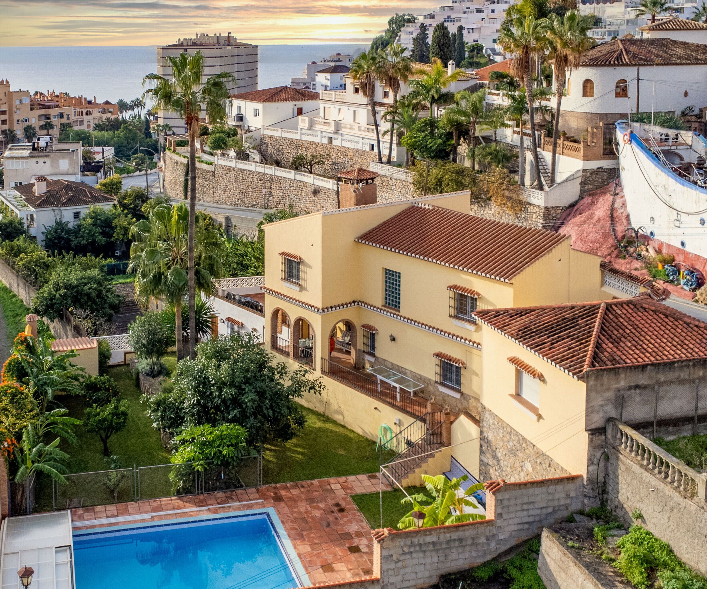 A Mediterranean-style villa with a swimming pool, surrounded by lush greenery and palm trees, overlooking a coastal town.