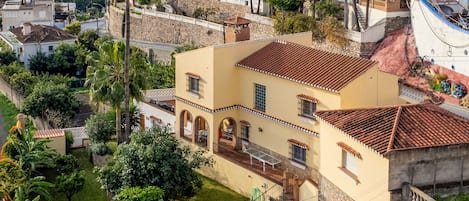 A Mediterranean-style villa with a swimming pool, surrounded by lush greenery and palm trees, overlooking a coastal town.