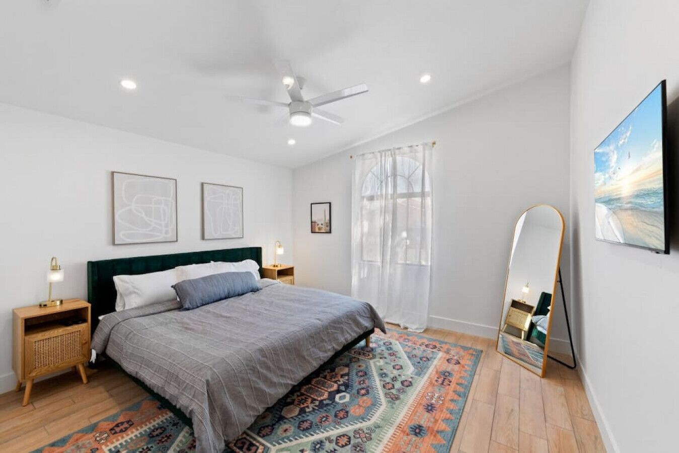 Ein modernes Schlafzimmer mit Bett, Deckenventilator, großem Spiegel und Blick ins Freie durch ein Fenster.