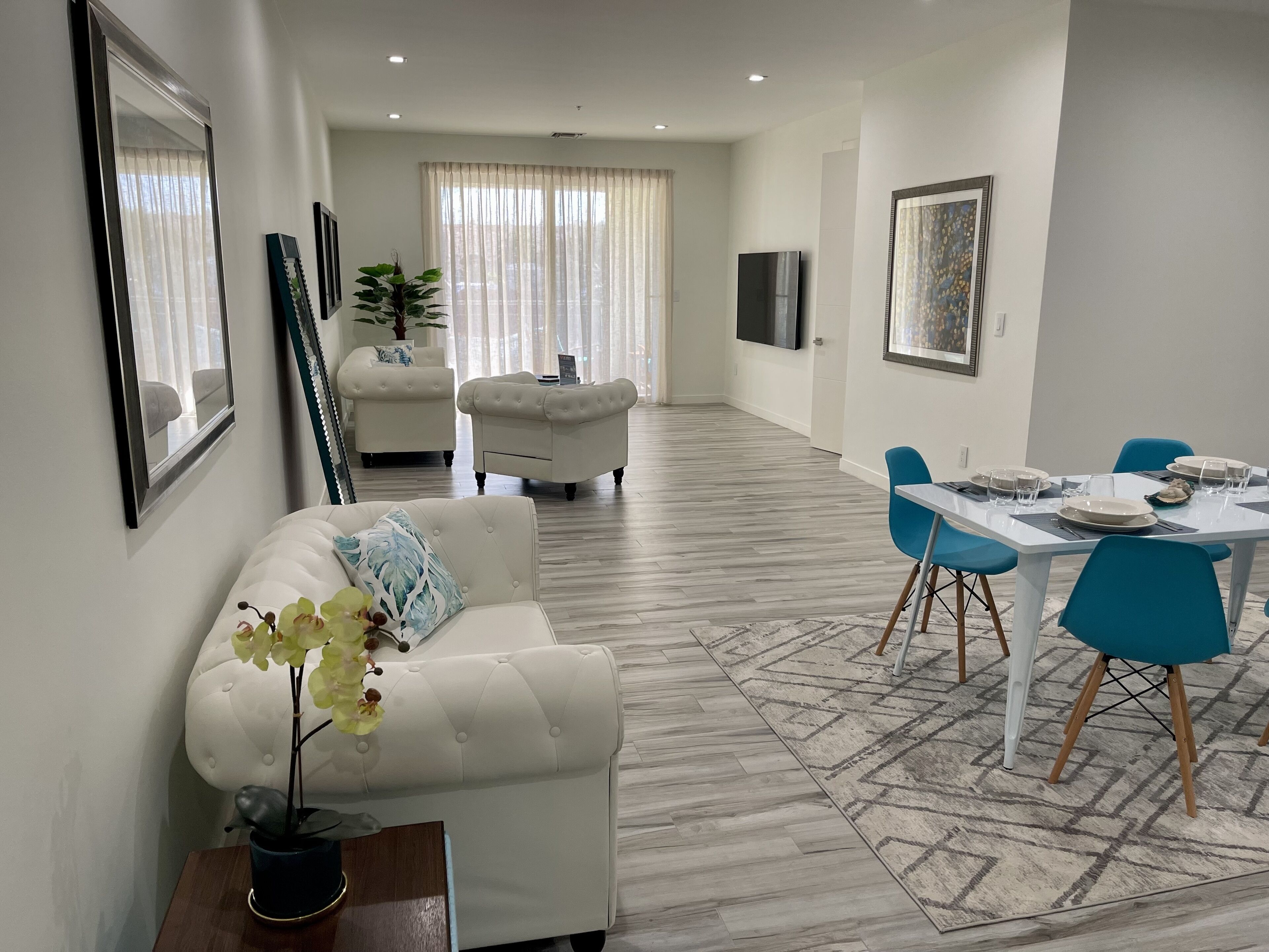 Una sala de estar moderna con un sofá blanco, una mesa de comedor con sillas azules y un ventanal con cortinas traslúcidas.