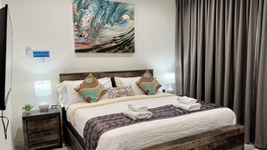 A neatly made bed with patterned bedding, a wooden headboard, and decorative pillows. There is a small bedside table with a lamp and a plant. A painting is hung above the bed, and a curtain is partially drawn to the side.