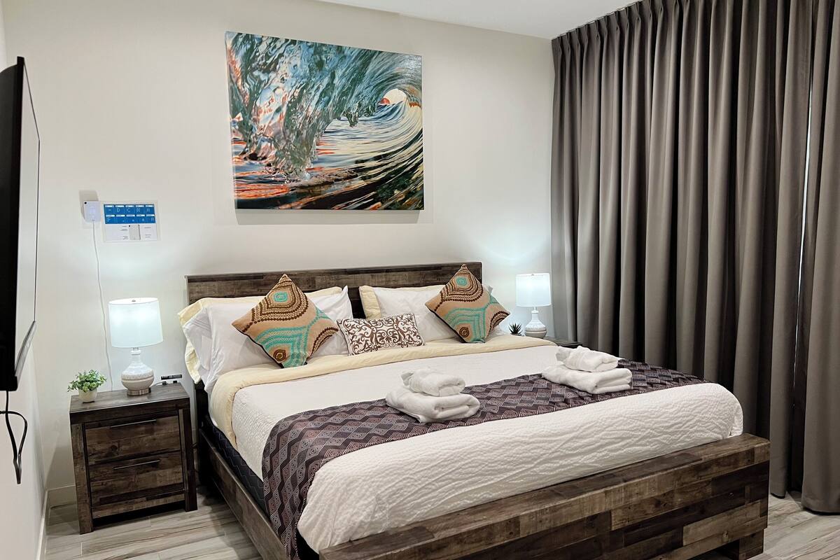 A neatly made bed with patterned bedding, a wooden headboard, and decorative pillows. There is a small bedside table with a lamp and a plant. A painting is hung above the bed, and a curtain is partially drawn to the side.