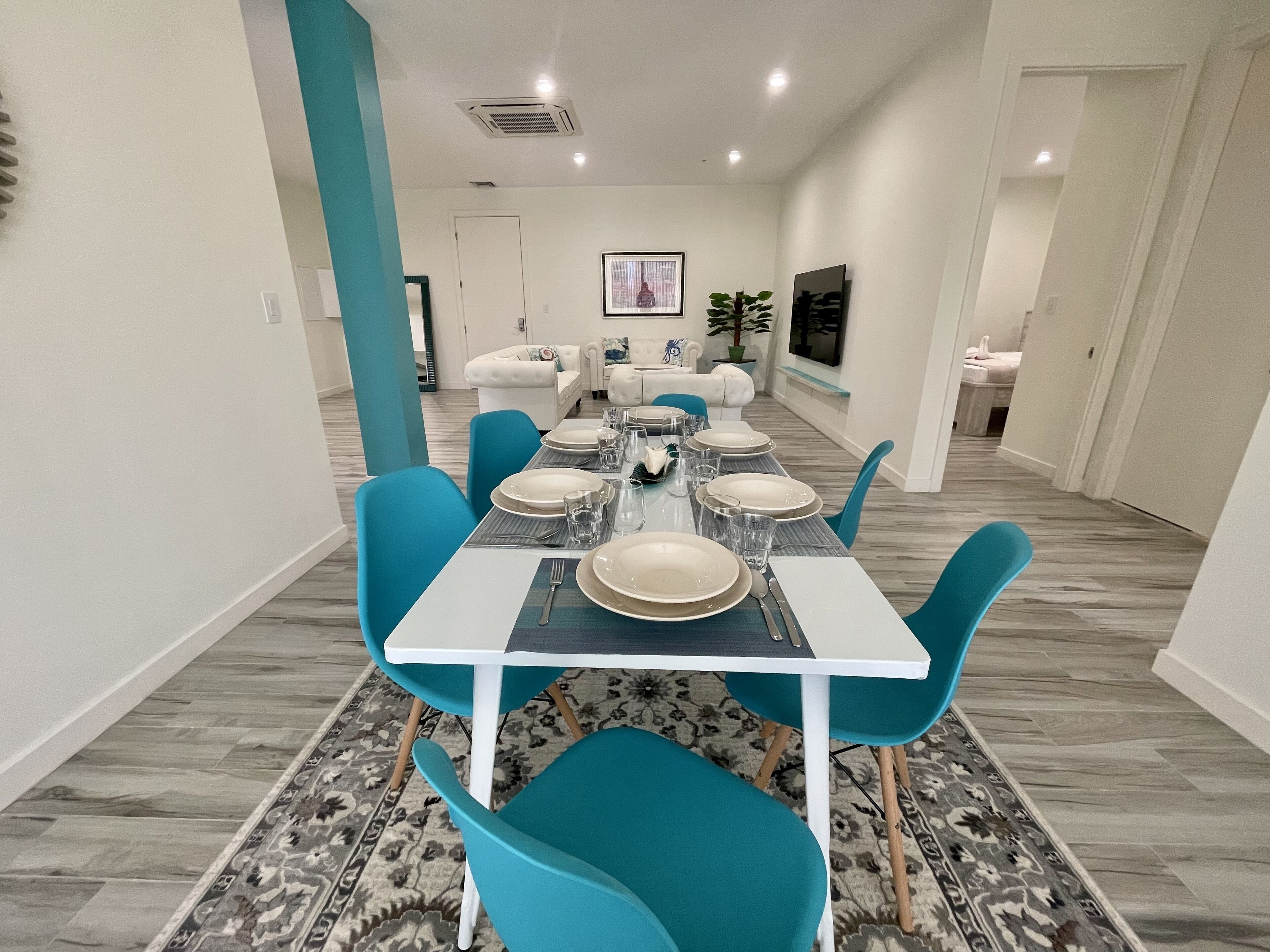 A dining area with a white table set for six, surrounded by teal chairs, a patterned rug, and a white sofa in the background.