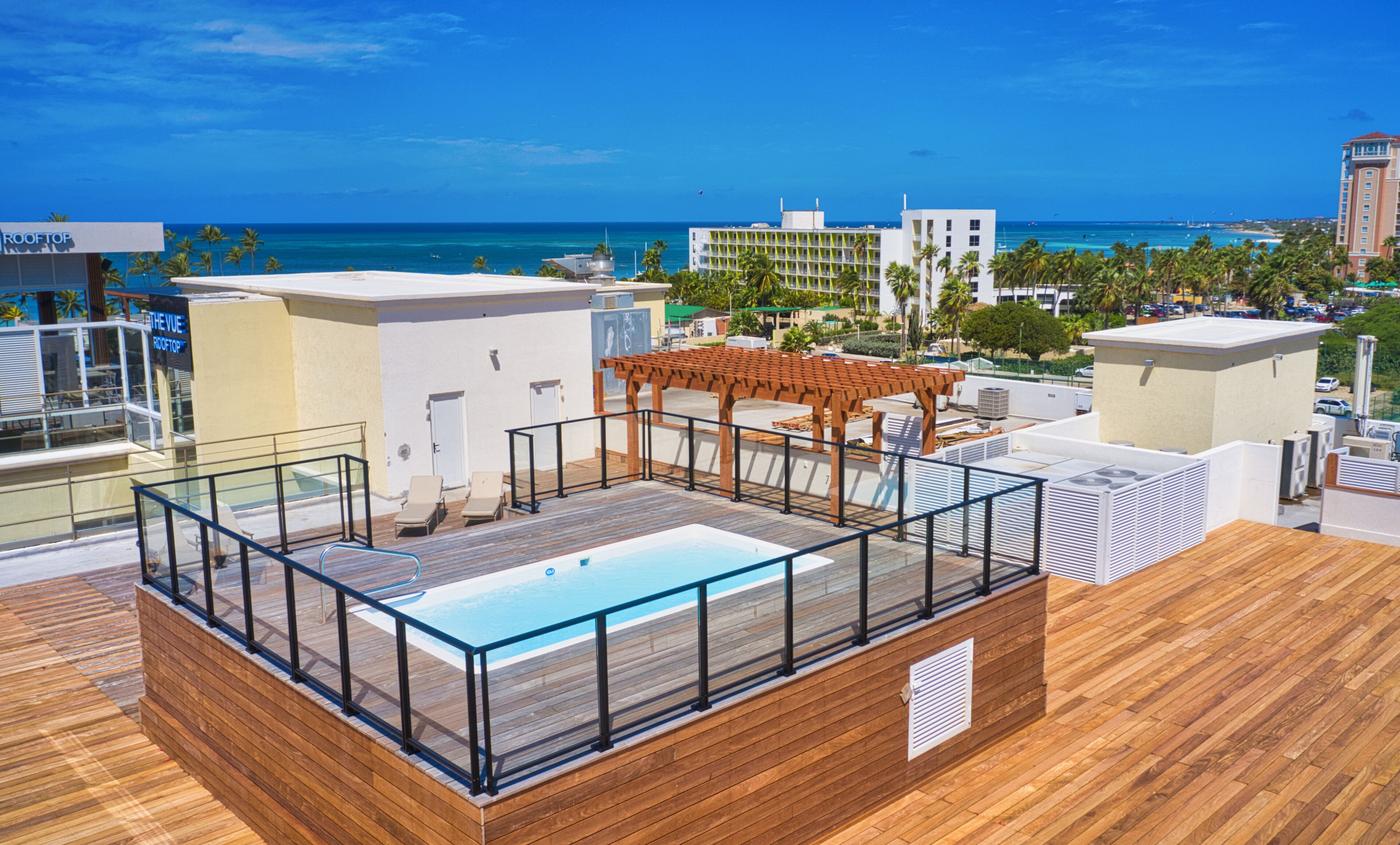 A rooftop pool area with a wooden deck, glass balustrade, and a pergola.