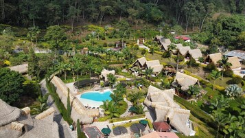 Un resort con piscina central, construcciones de techo de paja y abundante vegetación.