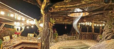 Un área de estar al aire libre con suelo de piedra, una estructura de árbol de madera y luces colgantes.