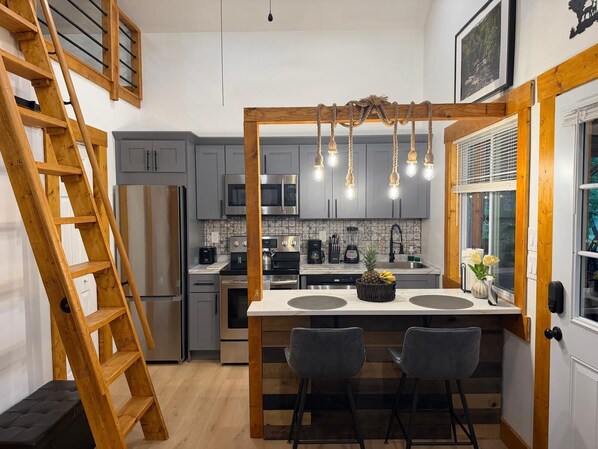 A modern kitchen with grey cabinets, a wooden ladder, a central island with two bar stools, and a hanging light fixture.